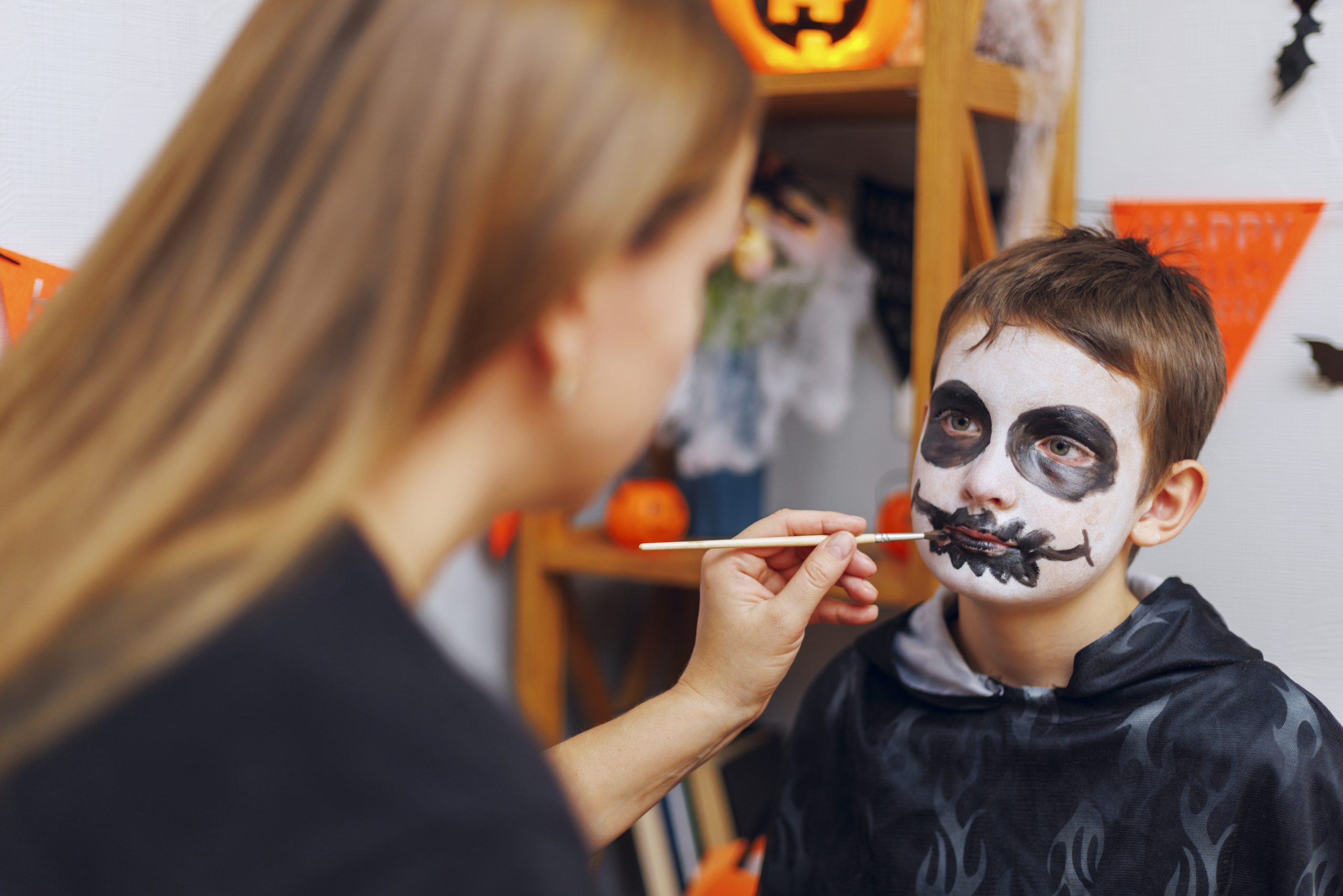 Woman applying skeleton face paint on a boy's face for Halloween celebration. The boy is dressed in a dark costume with Halloween decorations in the background